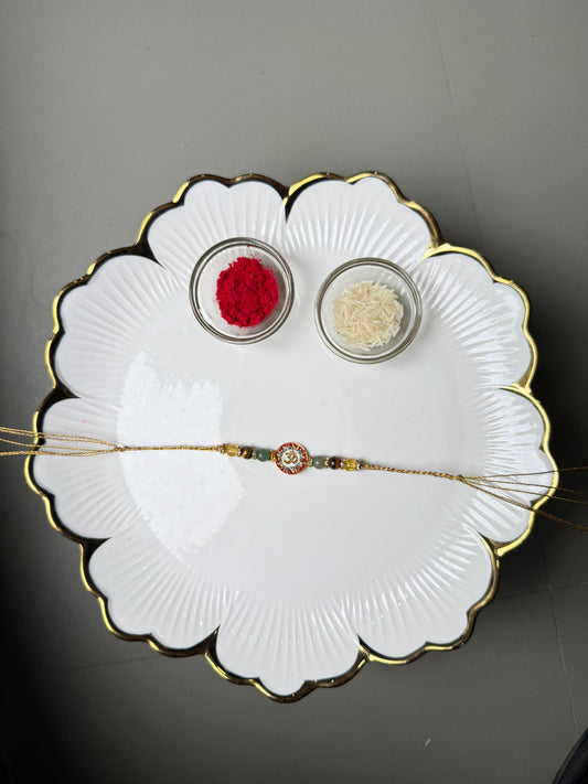 Decorative white plate with gold rim featuring a Rakhi, red and white powders, and a bracelet on a gray background.